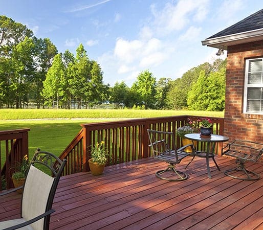 Cherry wood stained deck on a red brick house with a bistro set, flower pots and astairs leading to a green lawn surrounded by hedges and trees. 